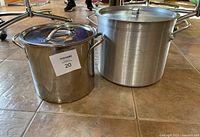 Two stock pots placed on tiled floor side by side showing size difference and handles, with lids on top.