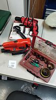 All three tools displayed on table: Black & Decker orange sander, red and black Hyper Tough hammer drill, and red Milwaukee hole saw kit case