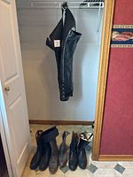 Black leather riding chaps hanging showing full length and boots on the floor below.