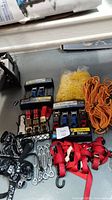 Photo of multiple ratchet straps with blue handles, red hook straps, orange rope, yellow plastic chain, and metal quick links spread on the floor.