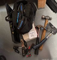 Overhead photo of black Mastercraft tool bag filled with various hand tools including screwdrivers, alongside two hammers and one hatchet with sheath.
