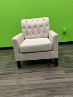 Front view of one beige upholstered armchair against green background, showing tufted backrest, armrests, wooden legs, and cushioned seat.