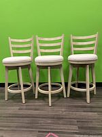 Front view of 3 matching wooden barstools with cream round cushion seats against green wall.