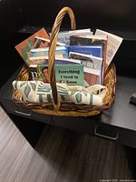 Wicker basket filled with various books and folded canvas bag inside, showing overall contents of lot.