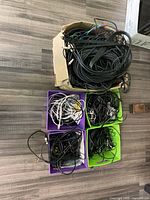 Top-down view of five containers (4 bins and 1 box) filled with various tangled cables of different colors including black and white cords, showing assortment of electronic cables and power cords.