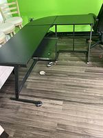 Full view of the L-shaped black desk showing keyboard shelf and caster wheels on steel frame.