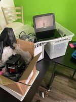 Phillips portable DVD player on top of plastic container labeled 'Video Computer', Polaroid camera in a cardboard box with bubble wrap, some other black fabric or cases visible