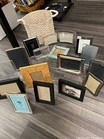 Photo showing 13 assorted small picture frames in different sizes, colors, and materials arranged upright on the floor around a cream woven basket with handles.
