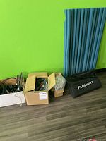 Wide shot of the lot showing three boxes/bags against a green wall and blue curtain: a white box with wires, a brown box with cables and hardware, and a black fabric bag labeled Floght.