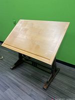 Front angled view of wooden drafting table with natural wood top and dark wooden base against green wall.