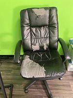 Front view of black office chair showing heavy wear and peeling on seat, backrest, and armrests against a green wall and wooden floor.