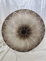 Front view of the round glass decorative plate showing an abstract flower with white petal outlines on a brown gradient and frosted glass surface.