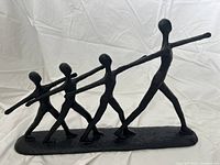 Front angle view of black plastic abstract statue showing four human figures in line, arms outstretched.