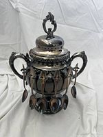 Front and side view of ornate silver plated sugar bowl with two handles, detailed lid, and twelve small spoons hanging on hooks around the bowl.