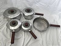 Full set of 5 Magnalite pots and pans with lids and wooden handles laid out on white cloth background.