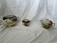 Three porcelain urns placed on white fabric backdrop showing floral, countryside designs and color variations.