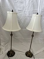Two 25 inch high brass table lamps with white linen bell-shaped shades and decorative ornate brass bases on a white cloth background.