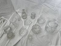 Photo showing the full collection of clear glass and crystal serving pieces arranged on white fabric background, highlighting the variety of shapes and sizes including bowls, pitcher, decanter, and covered jar.