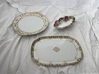Three assorted Nippon porcelain serving dishes positioned on white cloth: a large round platter, smaller rectangular platter, and floral hand-painted oval bowl