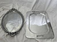 Wide view of both metal trays on white draped fabric background, showing oval tray with rope handle design and rectangular tray with cutout handles.