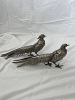 Pair of vintage silver metal bird ornaments placed side by side showing full body, tails, and feet details on white fabric background.