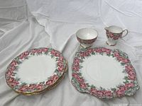 Group photo of 4 lunch plates, 1 serving plate, cream jug and sugar bowl all with white base, pink rose floral border and gold trim.
