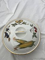 Top view of round porcelain serving dish with painted fruits and gold trim, showing central handle and glossy finish.