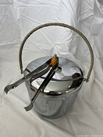 Side view of silver metal ice bucket with woven handle and metal ice tongs attached to lid