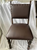 Front view of single dining chair showing leather upholstered seat and back, brass nailhead trim, carved legs