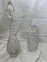 Two-piece vintage crystal decanter set with detailed cut glass patterns: one tall decanter with handle and oval stopper, and one smaller square decanter with geometric stopper.