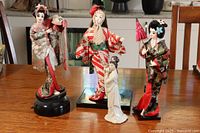 Photo showing four Asian dolls on a wooden table, three on stands wearing traditional kimonos, one smaller porcelain doll in white dress.