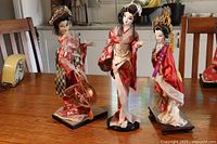 Photo of three Asian dolls standing on individual black stands, showing front and side views of dolls with detailed traditional costumes and ornate hair accessories