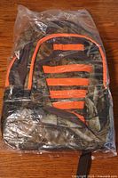 Front view of new camouflage backpack inside clear plastic packaging showing orange stripes and black mesh side pocket.