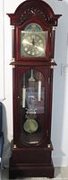 Full view of tall grandfather clock showing reddish mahogany finish, carved arch top, glass door with visible pendulum and chime rods inside.