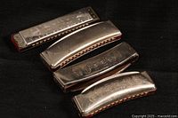 Four vintage harmonicas in line, showing tops and holes, metal bodies with engravings