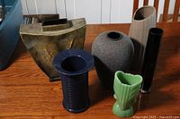 Six vases of different styles and materials including jadeite, ceramic, glass, and metal, displayed on a wooden table.