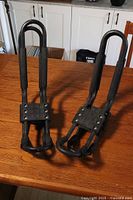 Two black metal roof rack supports with foam padding viewed from front, placed on wooden table