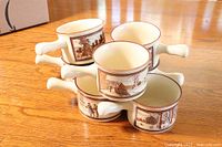 Six ceramic French onion soup bowls stacked, showing cream color and brown festive scenes on the sides.