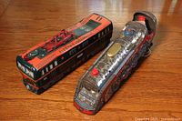 Two tin litho toy trains on a wooden surface: a red and black electric trolley bus and a silver and red steam locomotive with visible wear and paint loss.