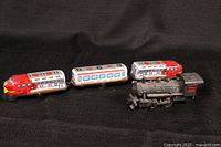 Two model trains displayed side by side on black fabric background, a three-piece colorful tin lithograph train and a black metal steam locomotive