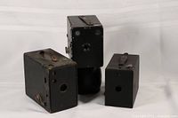 Three vintage box cameras arranged against a white background, showing front and side views. Cameras are black metal and leatherette with leather straps on two units. Wear and rust spots visible.