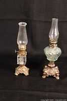 Two vintage oil lamps on ornate metal tripod bases with glass chimneys, one clear and one green glass reservoir.