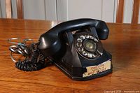 Black vintage rotary dial phone placed on wood surface with coiled cord and sticker reading 'CAUTION THIS LINE IS NOT SECURE'.