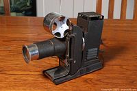 Side view of vintage 35mm film projector showing large lens barrel and metal reel arm on top, mounted on a wooden surface.