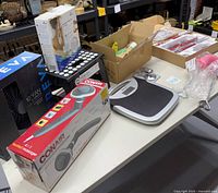 Photo of lot items on table showing boxed Treva 10 inch fan, Conair heated massager box, digital weight scale, shoe stretchers, dusters, and swivel sweeper