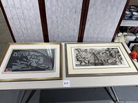 Two framed and matted woodblock prints on a white table with white and brown room dividers in the background. Left print has Hebrew text and dark abstract imagery; right print depicts figures around a chessboard.