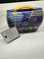 Front view of the limited edition Jetsons metal lunchbox with original tag attached showing cartoon characters, mainly in blue and yellow with a yellow handle.