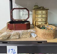 Photo of two jewelry boxes, wooden with glass top and gold painted wood with drawers, along with basket of beaded necklaces, vintage pearls in plastic bags, and other jewelry on shelf.