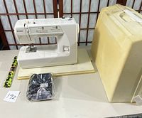 White Kenmore sewing machine on table with black power cord and foot pedal in plastic, yellow hard carrying case, and small green craft fabric strip with black cat silhouettes.