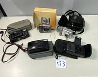 Five vintage cameras displayed on table including Canon digital cameras, Sears FF33 film camera, Yashica and Kodak film cameras, some with cases and strap.